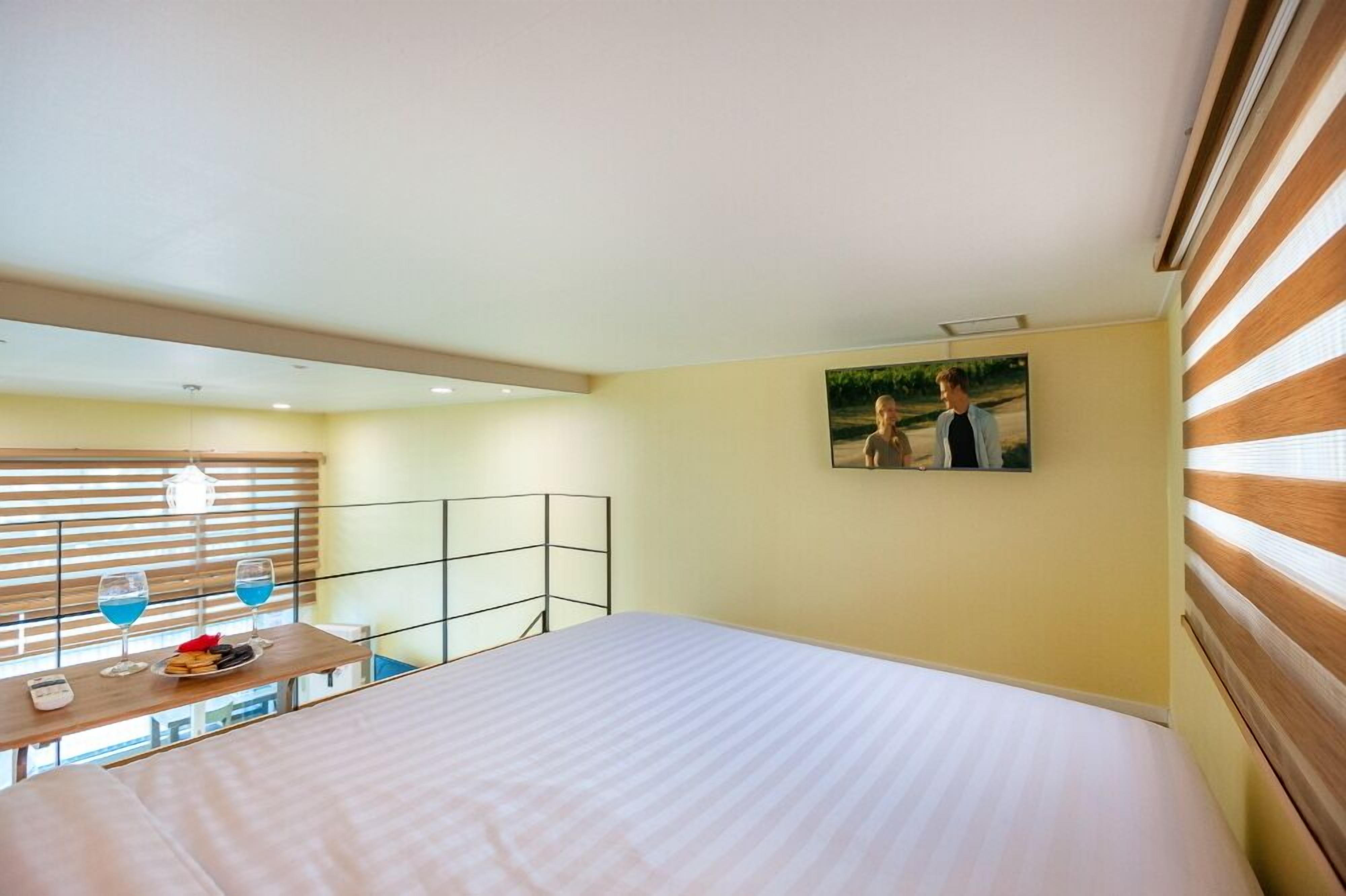 A hotel room with a bed, a small table with two glasses of blue liquid, a plate with snacks, a picture on the wall, and a window with blinds.