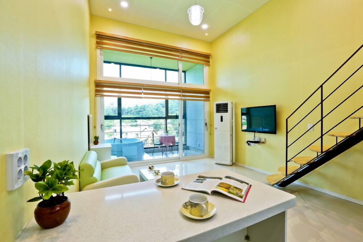 A bright room with a large window, a flat-screen TV, a table with cups and a book, and a staircase.