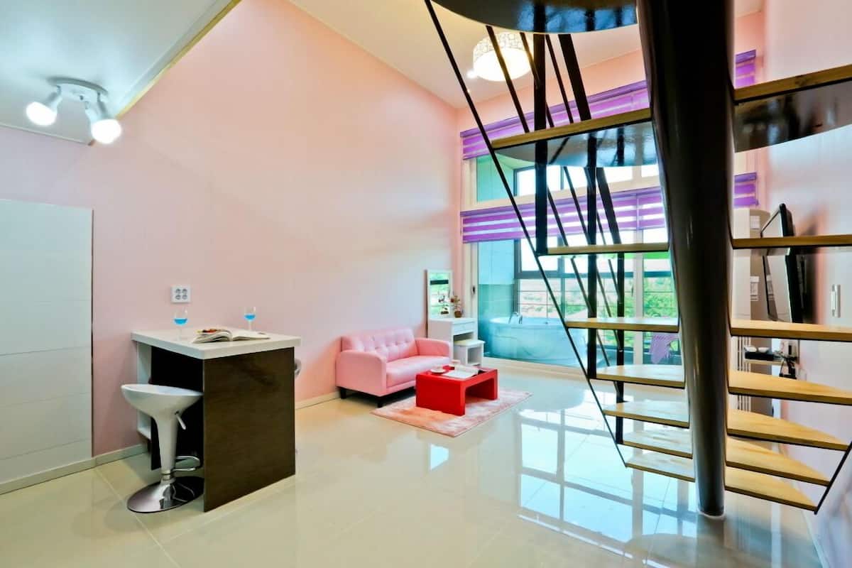 A modern interior with a pink sofa, a red coffee table, a white bar area, and a staircase with a glass railing.
