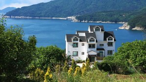 A two-story house with a gabled roof, located near a body of water with a mountainous backdrop. - Mulbit Hanul (Geoje)