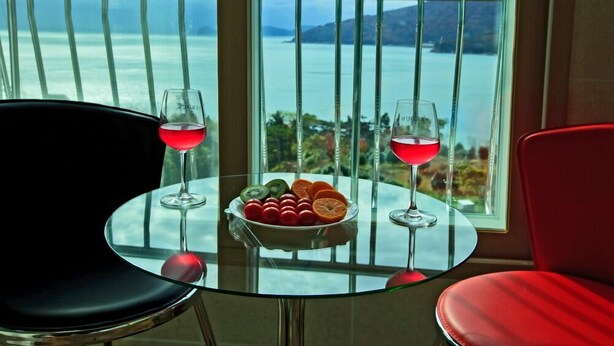A table with two glasses of red wine and a bowl of fruit in front of a window with a view of the sea and mountains. - Mulbit Hanul (Geoje)