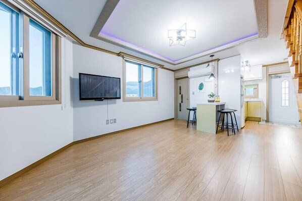 A spacious living area with a large TV, wooden flooring, and a kitchen area with stools.