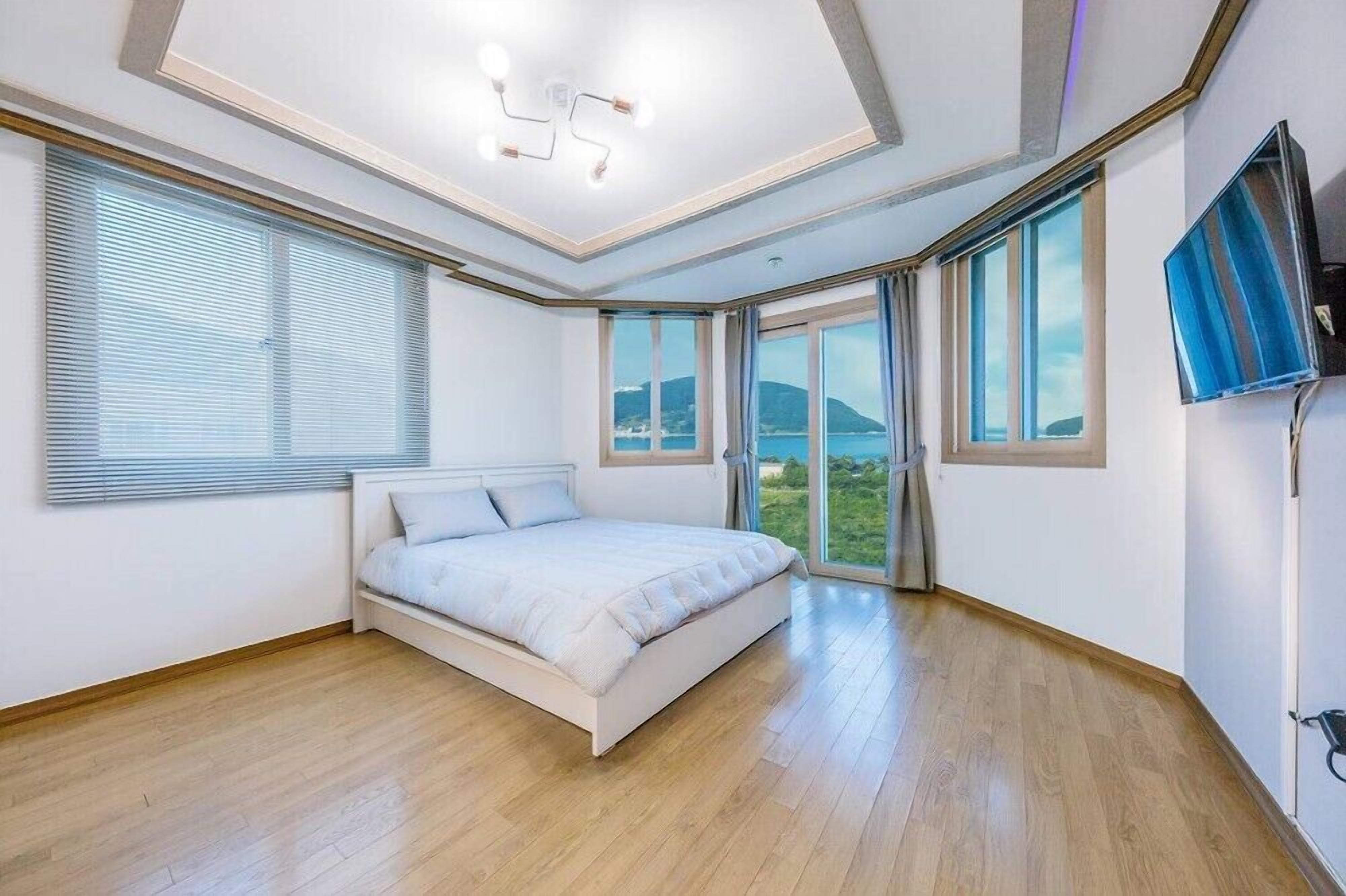 A bedroom with a bed, large windows, a TV, and a view of the sea.