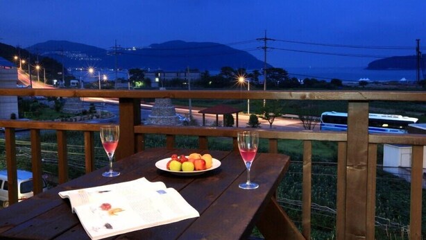 Una terraza de madera con una mesa preparada para dos personas, que incluye un plato de fruta y dos copas de vino tinto, con vistas a una zona costera y una montaña al fondo.