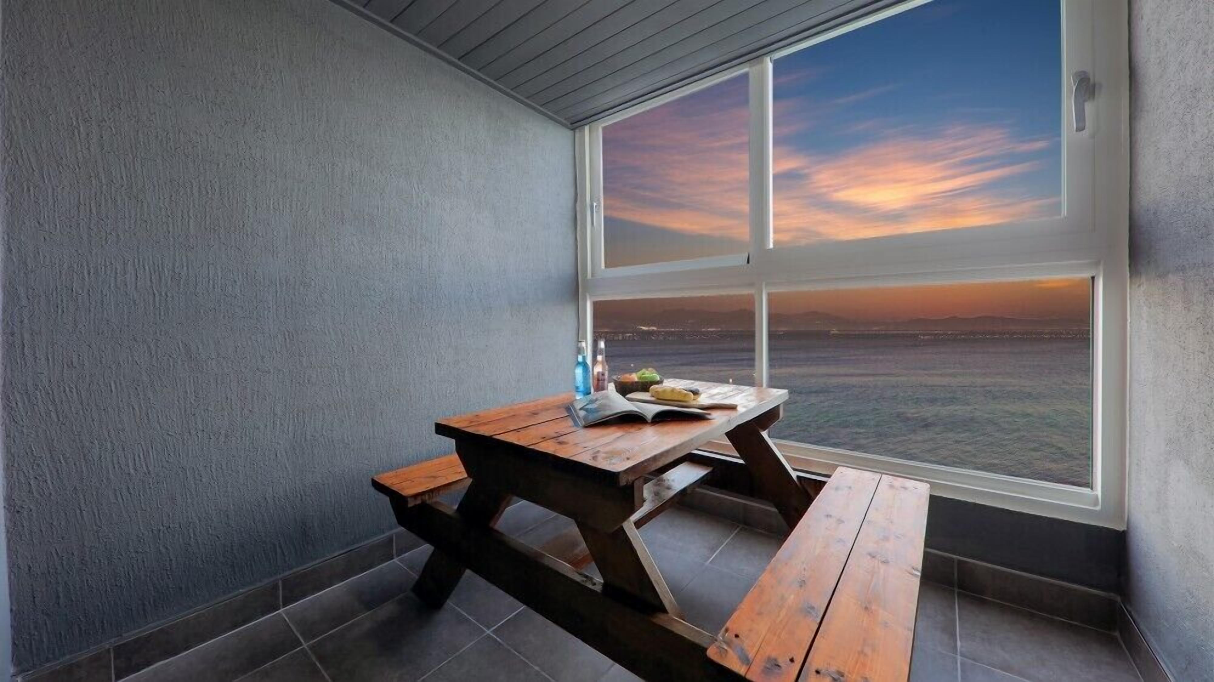 Un pequeño balcón cerrado con una mesa y bancos de madera, con vista a la puesta de sol sobre el océano.