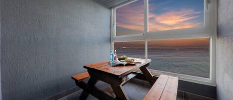 Un pequeño balcón cerrado con una mesa y bancos de madera, con vista a la puesta de sol sobre el océano.