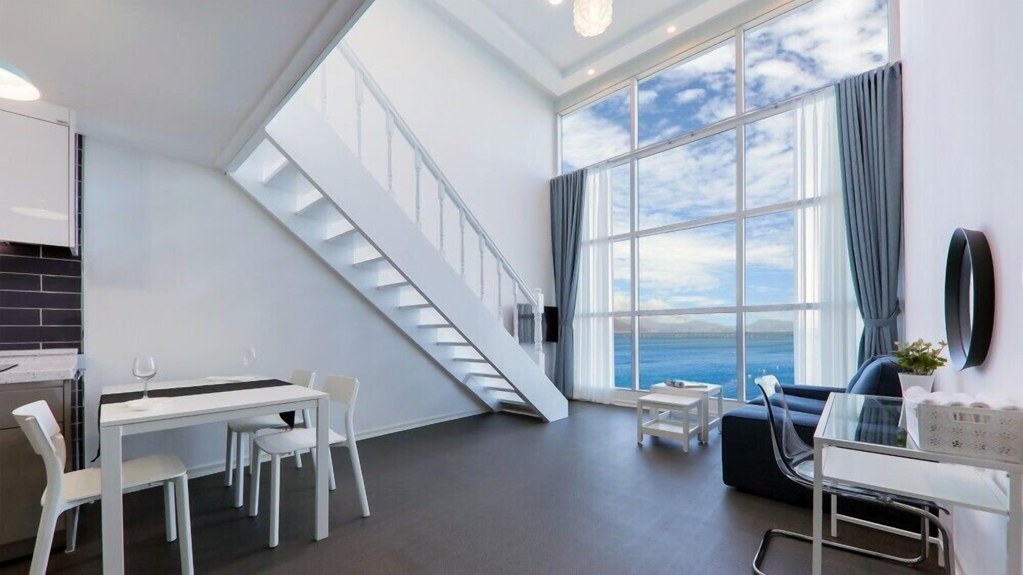 Una habitación moderna con comedor, una escalera y un amplio ventanal con vista al mar.