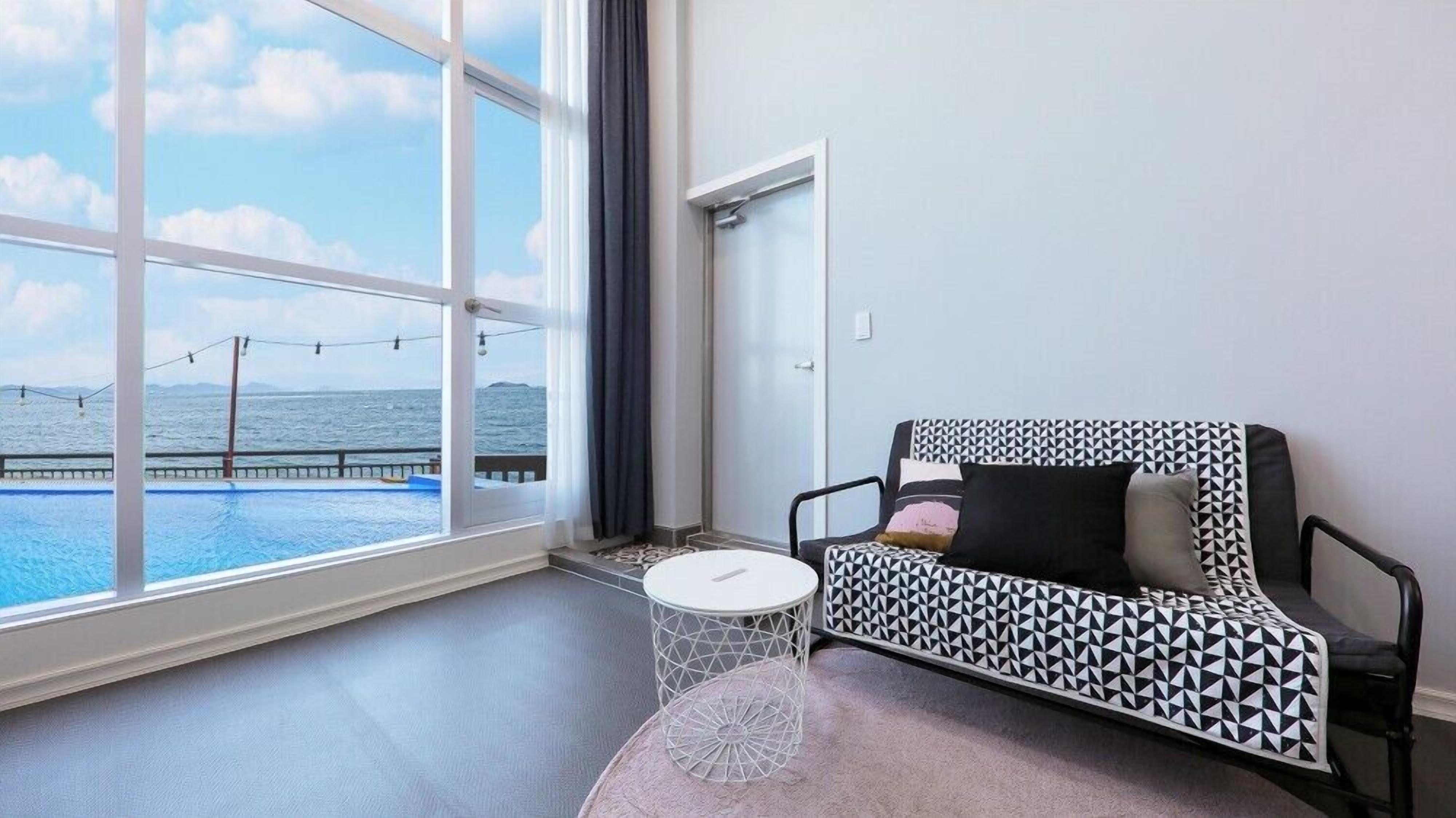 Una habitación moderna con un amplio ventanal con vistas a una piscina y al mar, un sofá blanco y negro estampado y una mesita redonda.