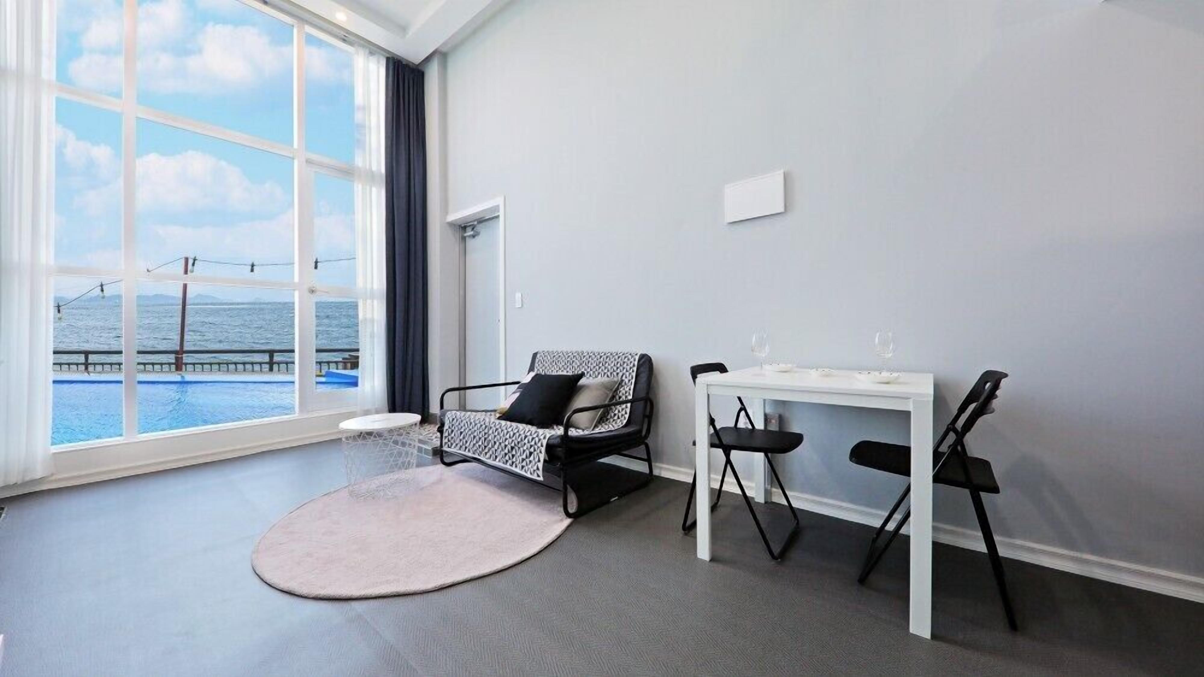 Una habitación moderna con un ventanal grande con vista a una piscina y al océano, una mesa blanca con dos sillas y un sillón estampado.