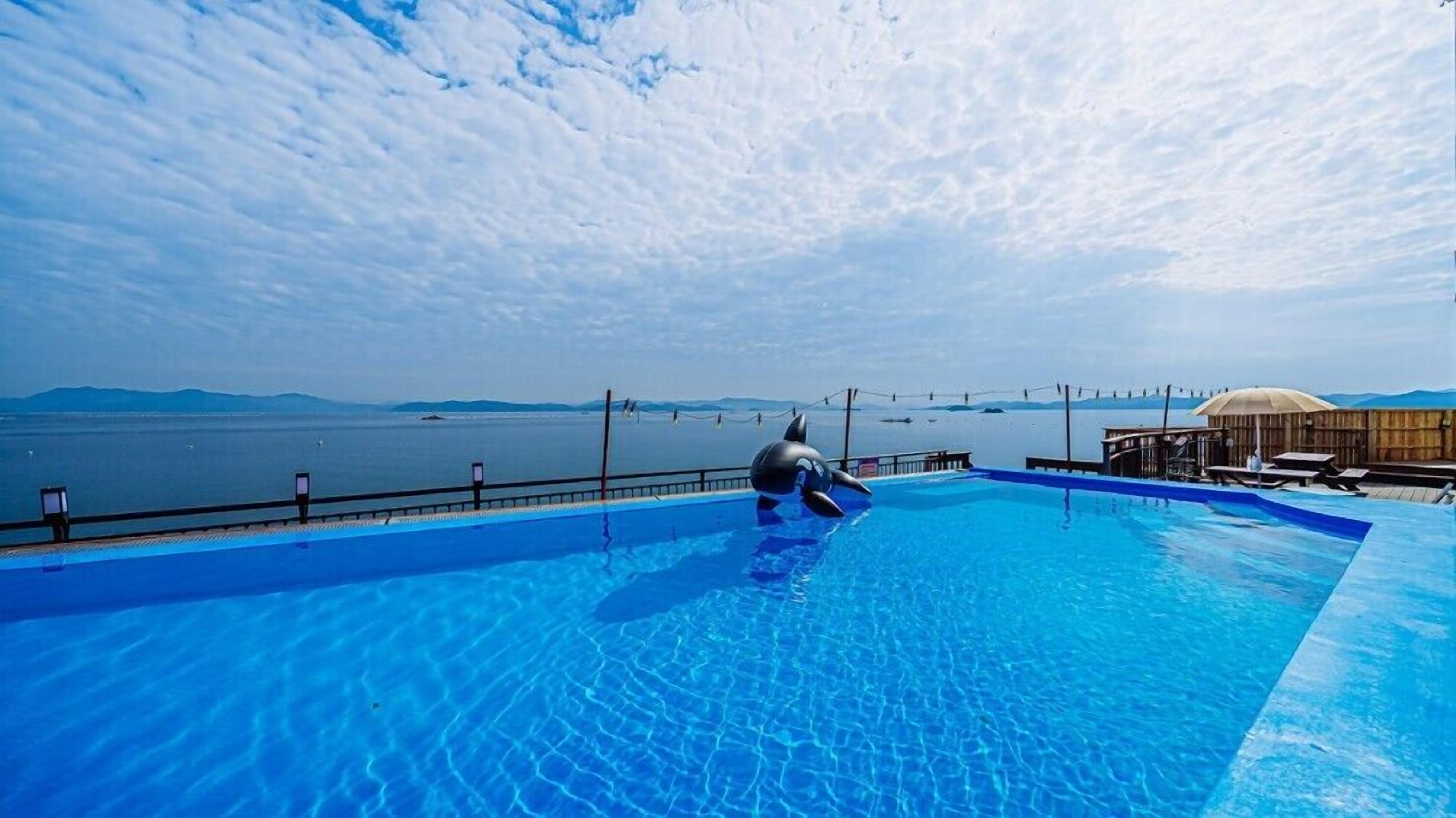 Ein Schwimmbecken mit einer Rettungswache, einer Holzterrasse mit Sonnenschirmen und Blick auf das Meer und die Berge.