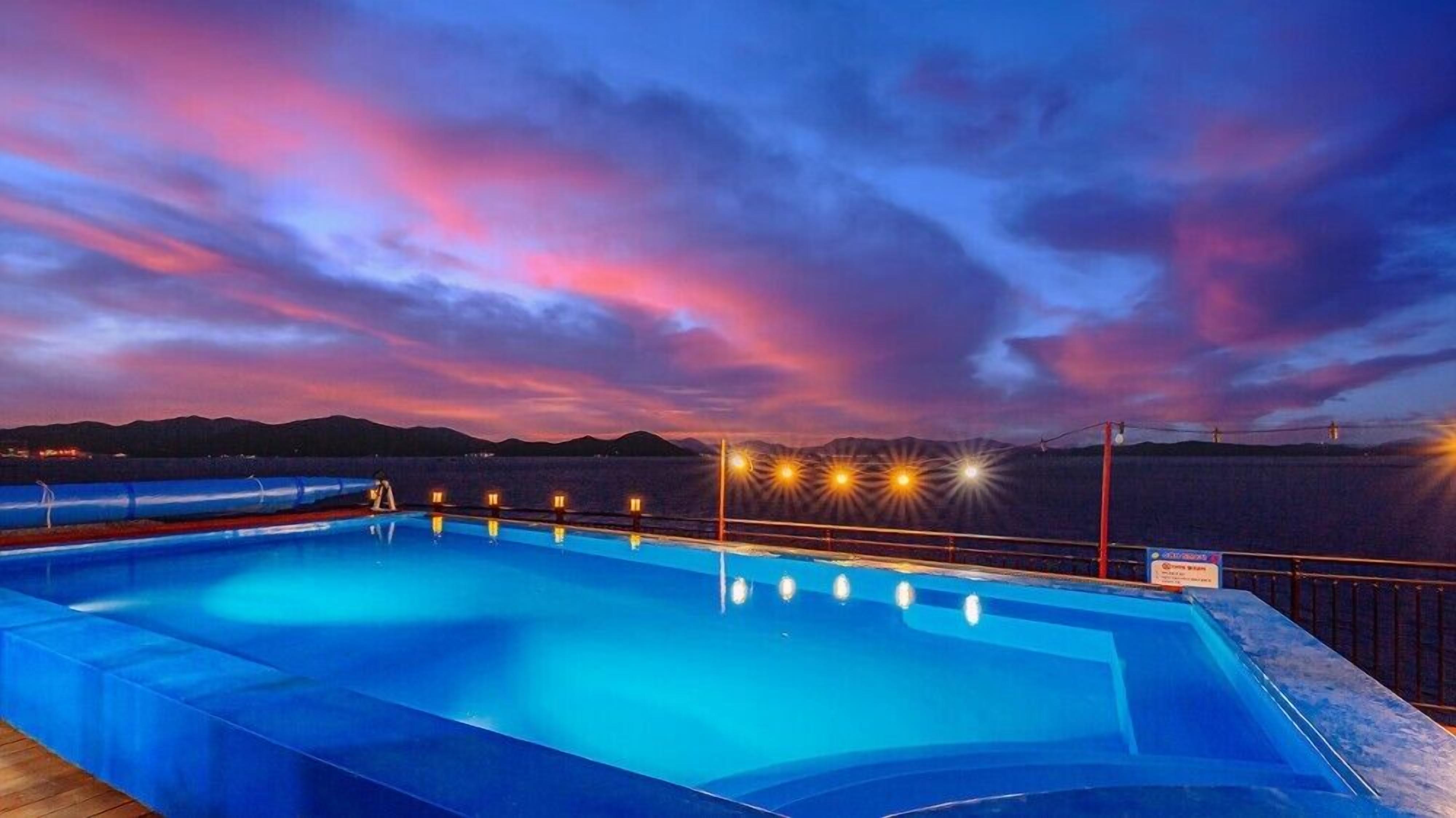 Una piscina al atardecer con cielo rojizo, rodeada de montañas e iluminada.