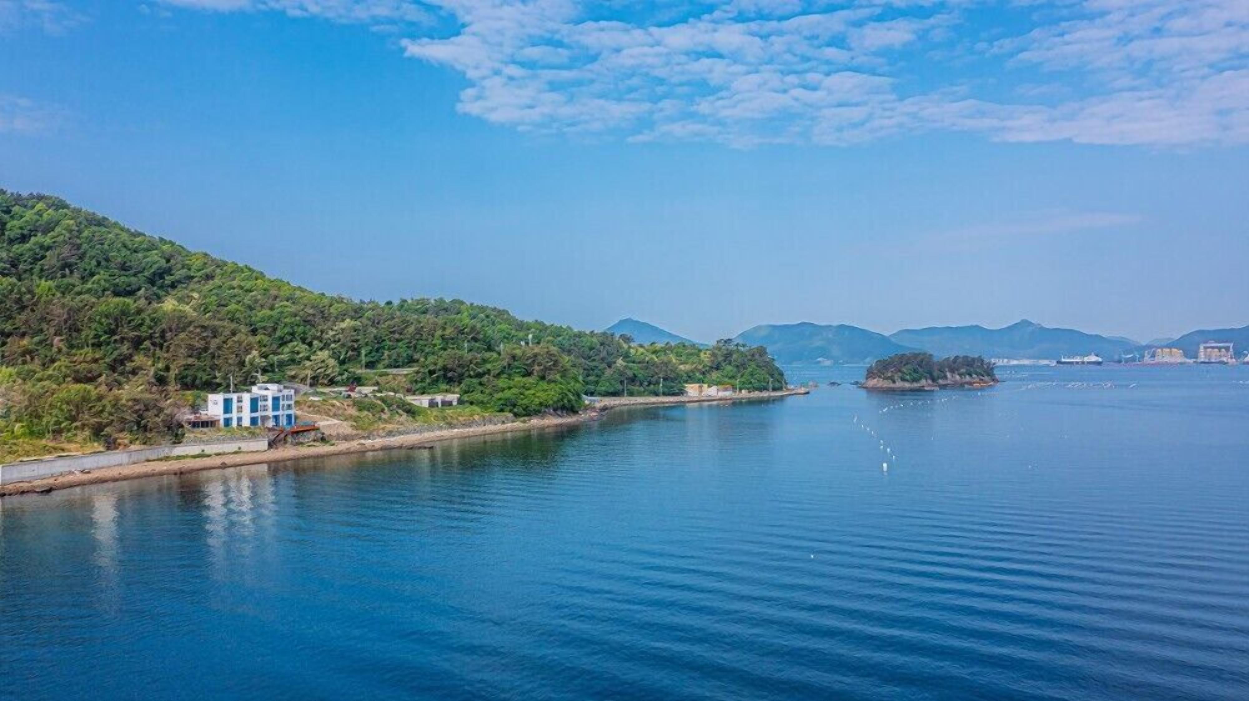 A coastal view with a building near the water, surrounded by hills and a clear sky.