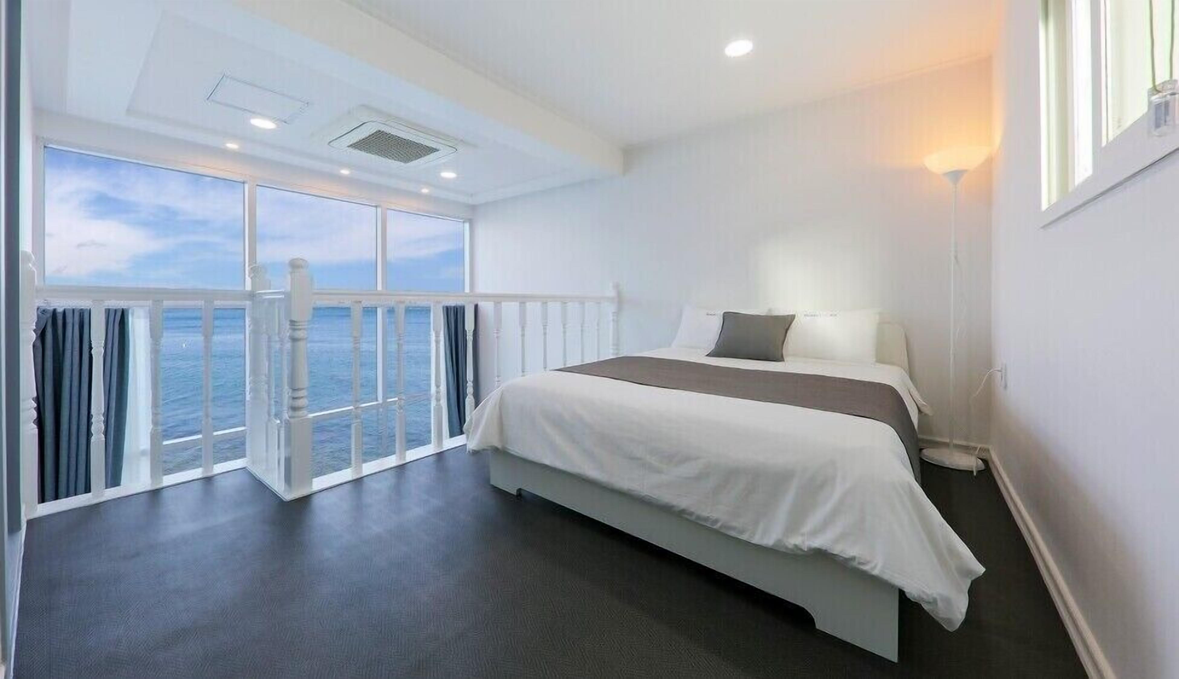A hotel room with a large bed, a view of the sea through floor-to-ceiling windows, and a balcony with a railing.