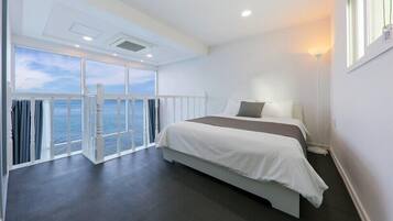 A hotel room with a large bed, a view of the sea through floor-to-ceiling windows, and a balcony with a railing.