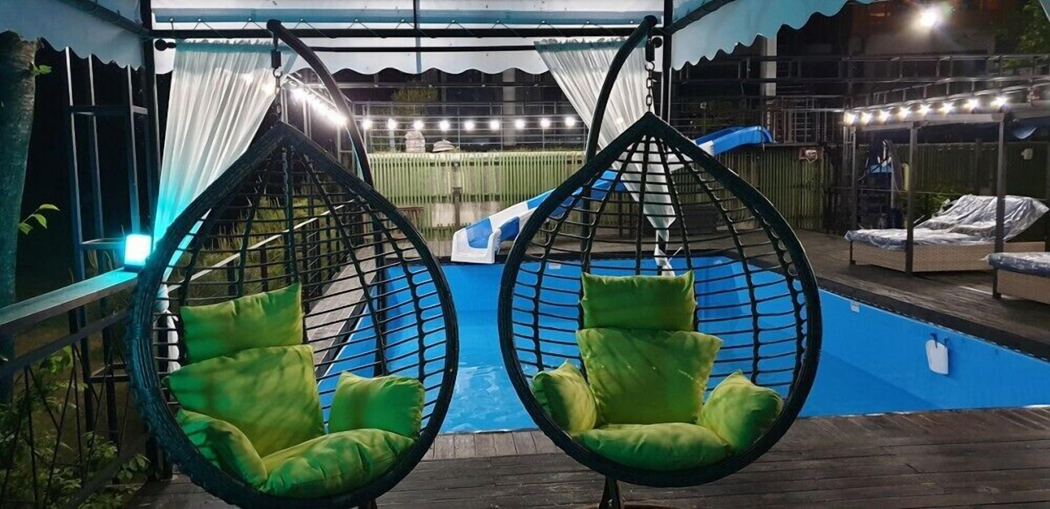 Two green hanging chairs by a pool with a covered area and a fence in the background.