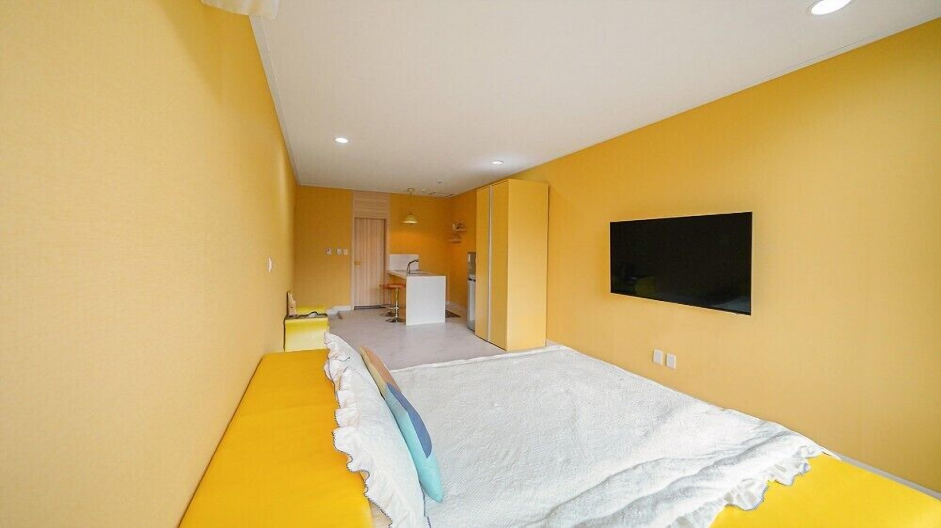 A narrow room with yellow walls, a bed with a white and blue blanket, a small kitchenette, and a flat-screen TV mounted on the wall.
