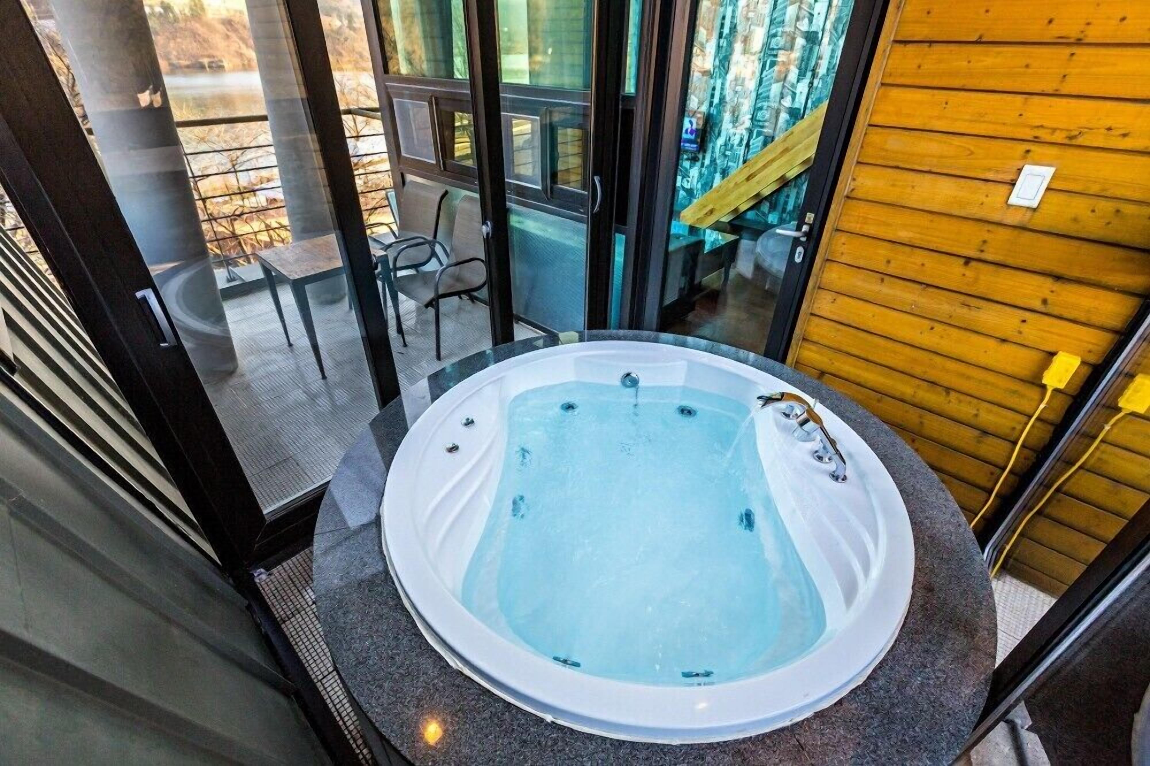 A hot tub in a room with a glass wall, a wooden wall, and a chair.