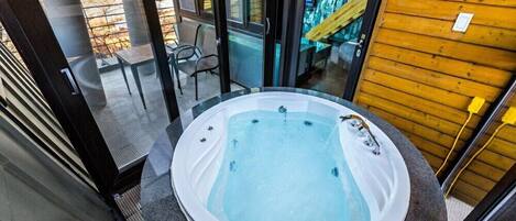 A hot tub in a room with a glass wall, a wooden wall, and a chair.