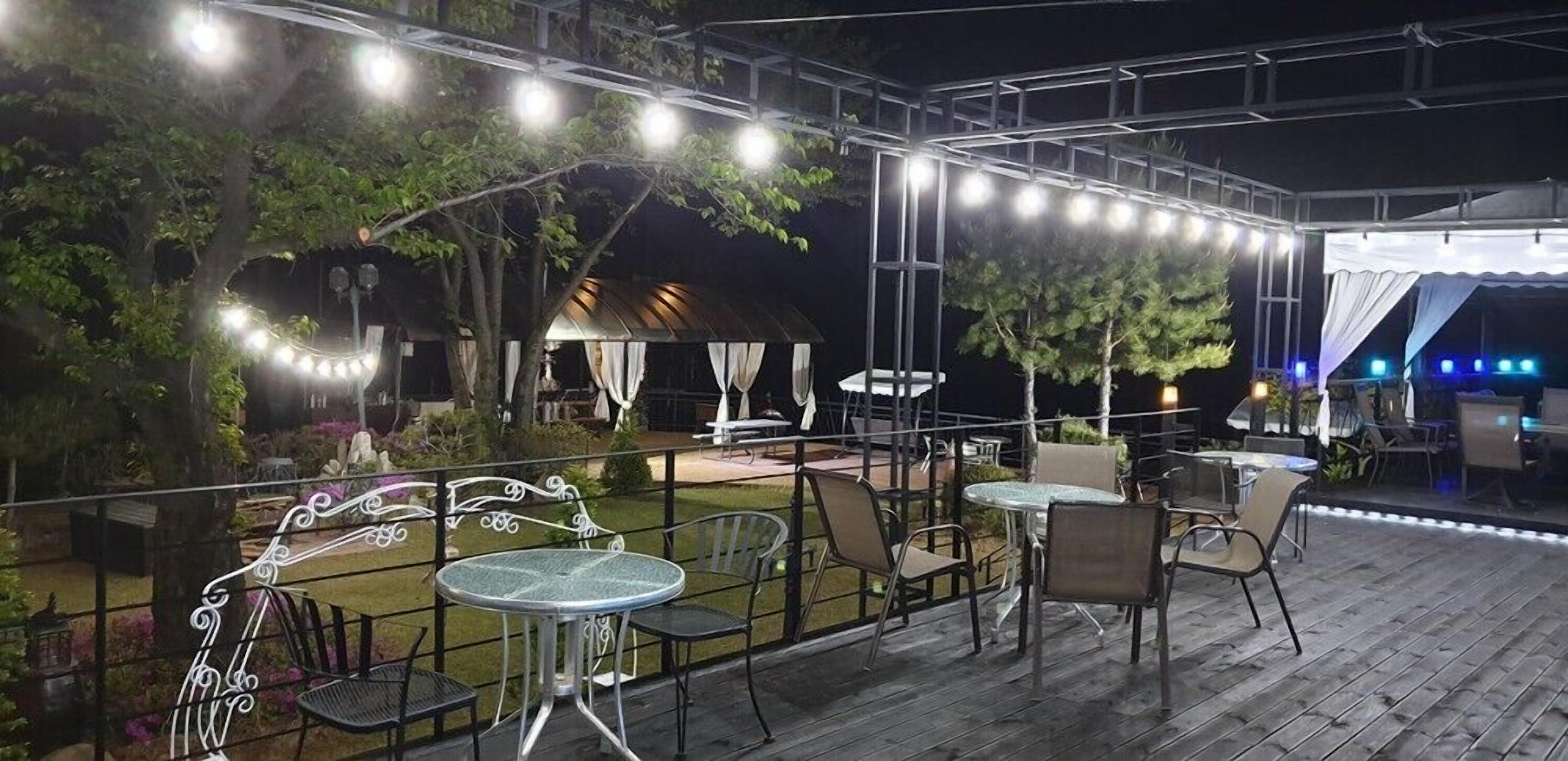 Un patio al aire libre con mesas y sillas, rodeado de árboles y una zona cubierta con luces colgantes.