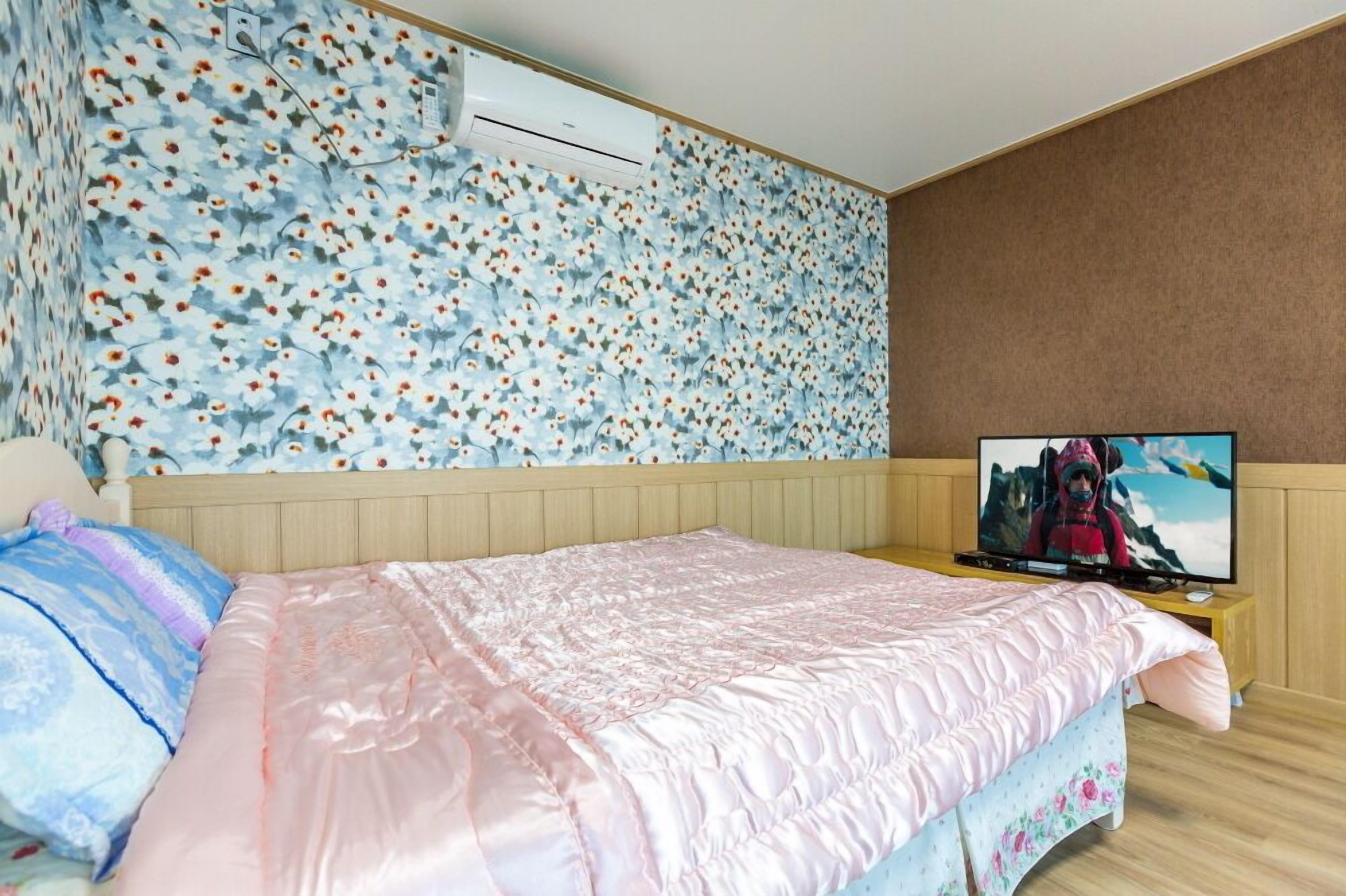 Un dormitorio con papel tapiz floral, una cama con ropa de cama rosa, un televisor sobre un soporte de madera y un equipo de aire acondicionado en la pared.