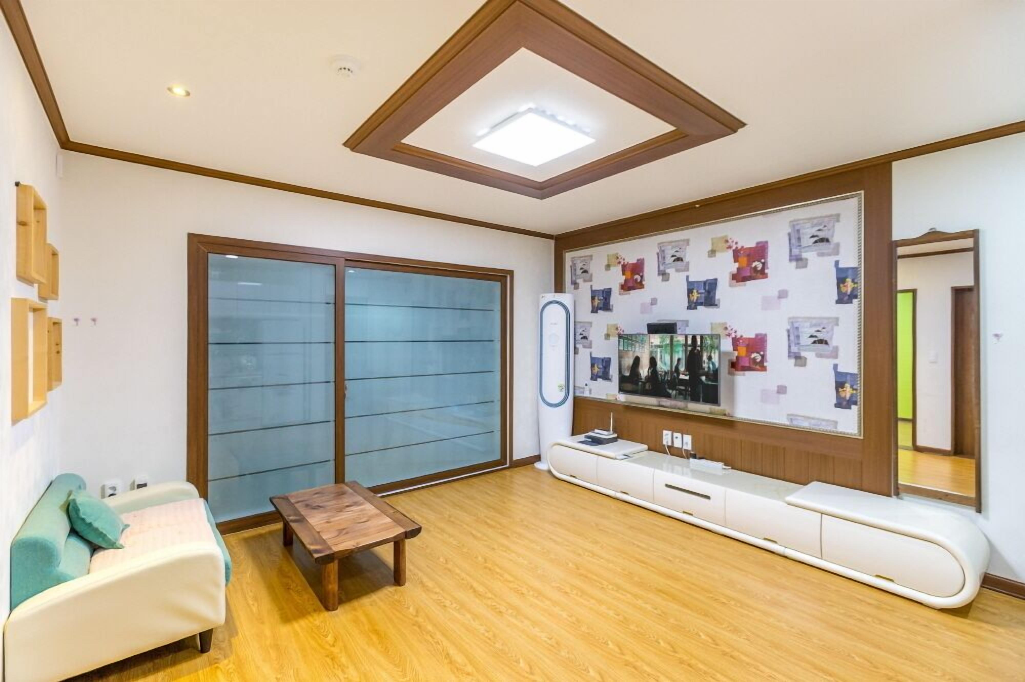 Una habitación con puerta corrediza de vidrio, un banco de madera, un sofá blanco, una mesa de madera, un televisor de pared y un espejo.
