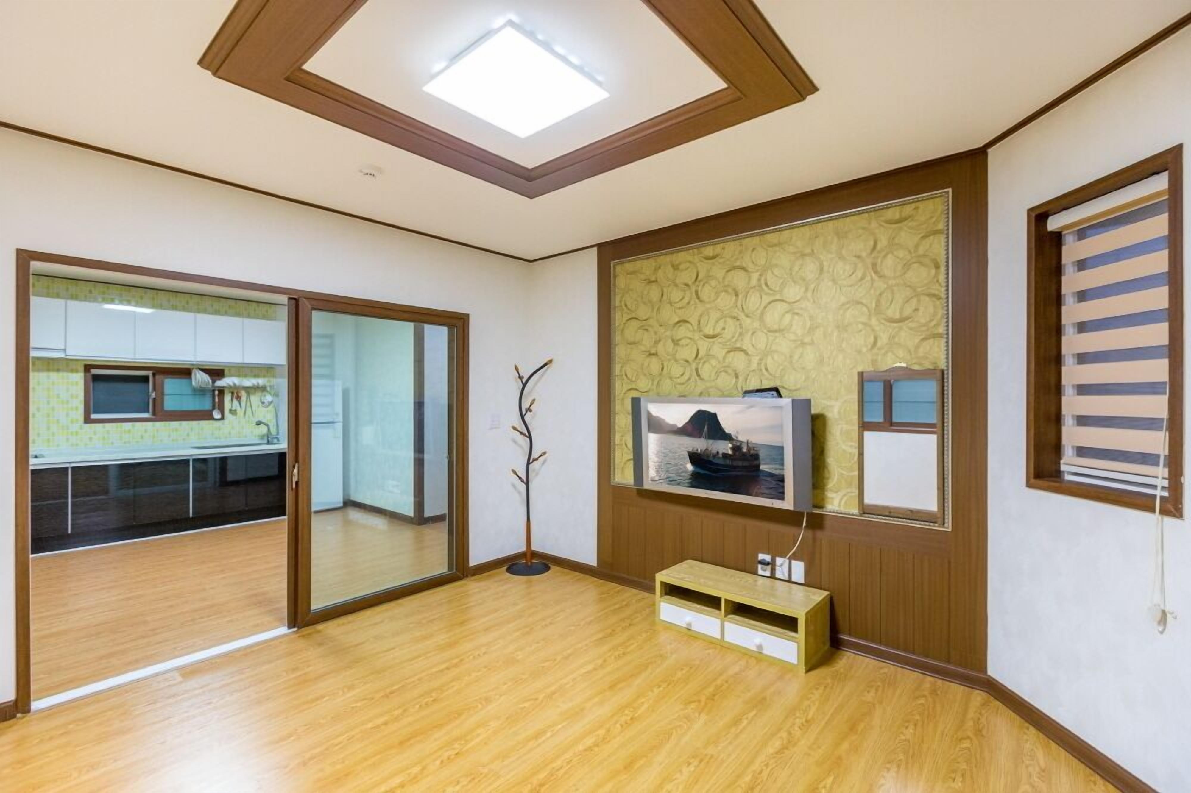Una habitación con piso de madera, una lámpara en el techo, un televisor montado en una pared con diseño y una puerta corrediza que da a una cocina.