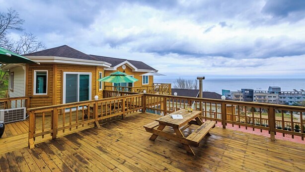 A wooden deck with a picnic table and umbrella, overlooking a body of water and buildings. - Neulpureun Pension (Yeongdeok)