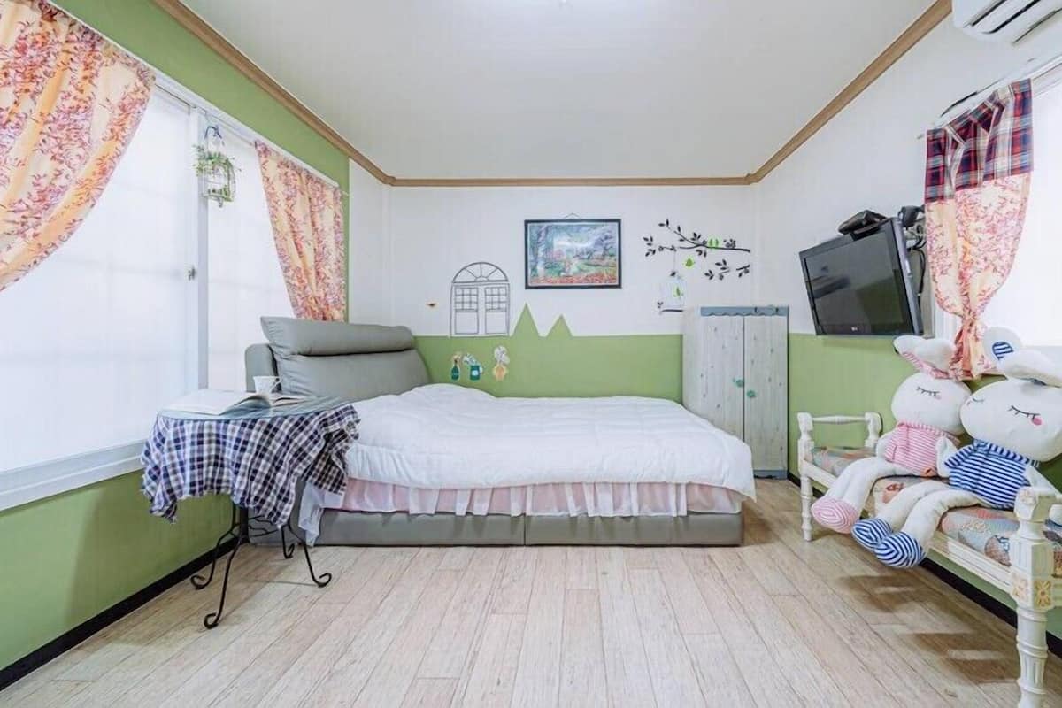 A bedroom with a bed, a small table, a chair, a TV, and a window with curtains.