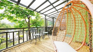 A wooden deck with a wicker swing chair and metal chairs, overlooking a green landscape. - Mulga Pension (Gapyeong)