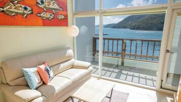 A balcony with a view of the sea and a mountain, a beige sofa with pillows, and a painting of fish.