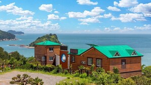 A wooden cabin with a green roof, surrounded by lush greenery and overlooking a coastal area with a clear sky. - Pado Hyanggi Pension (Geoje)