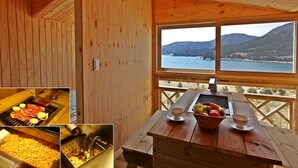 A wooden cabin with a dining area featuring a grill, a table with a basket of fruit, and a view of a lake and mountains. - Pado Hyanggi Pension (Geoje)