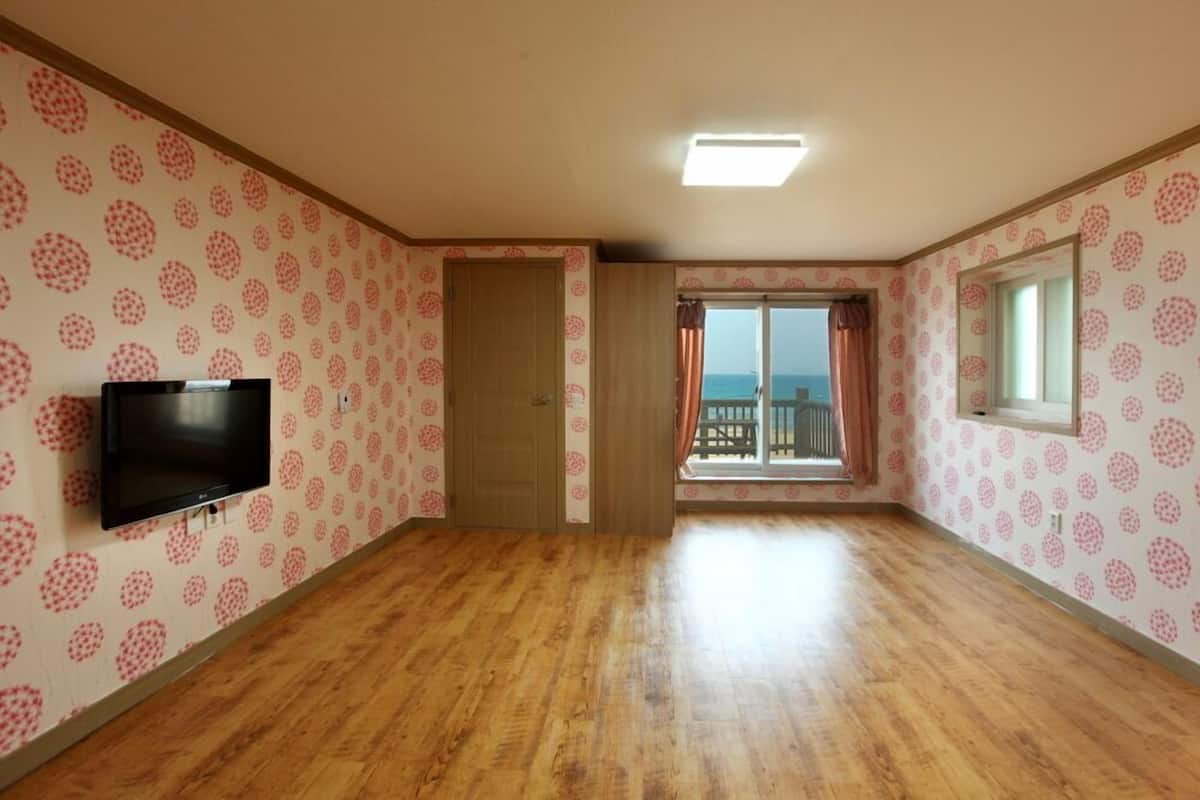 A room with a wooden floor, a television mounted on the wall, a door, and a window with a view of the sea.