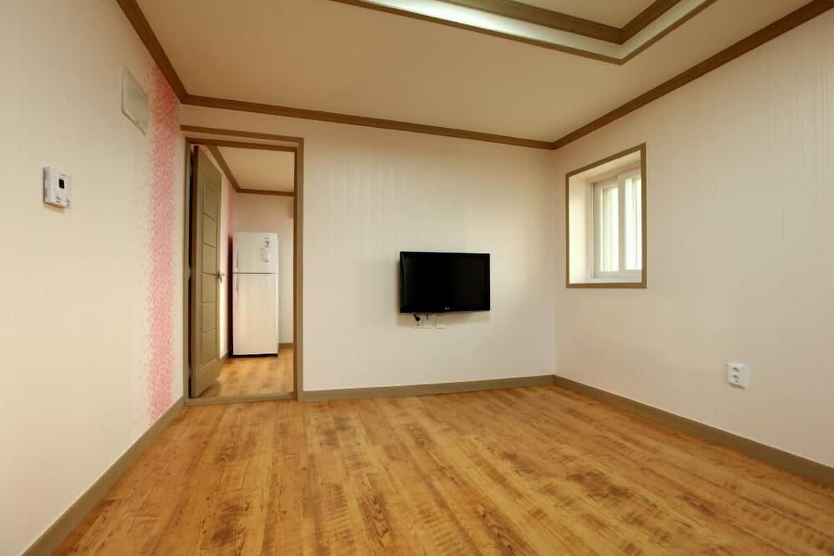 A room with wooden flooring, a flat-screen TV mounted on the wall, and a window providing natural light.