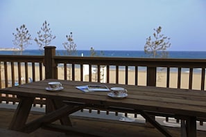 Una mesa de picnic de madera con dos tazas y una revista encima, con vistas a una playa con árboles y el océano al fondo.