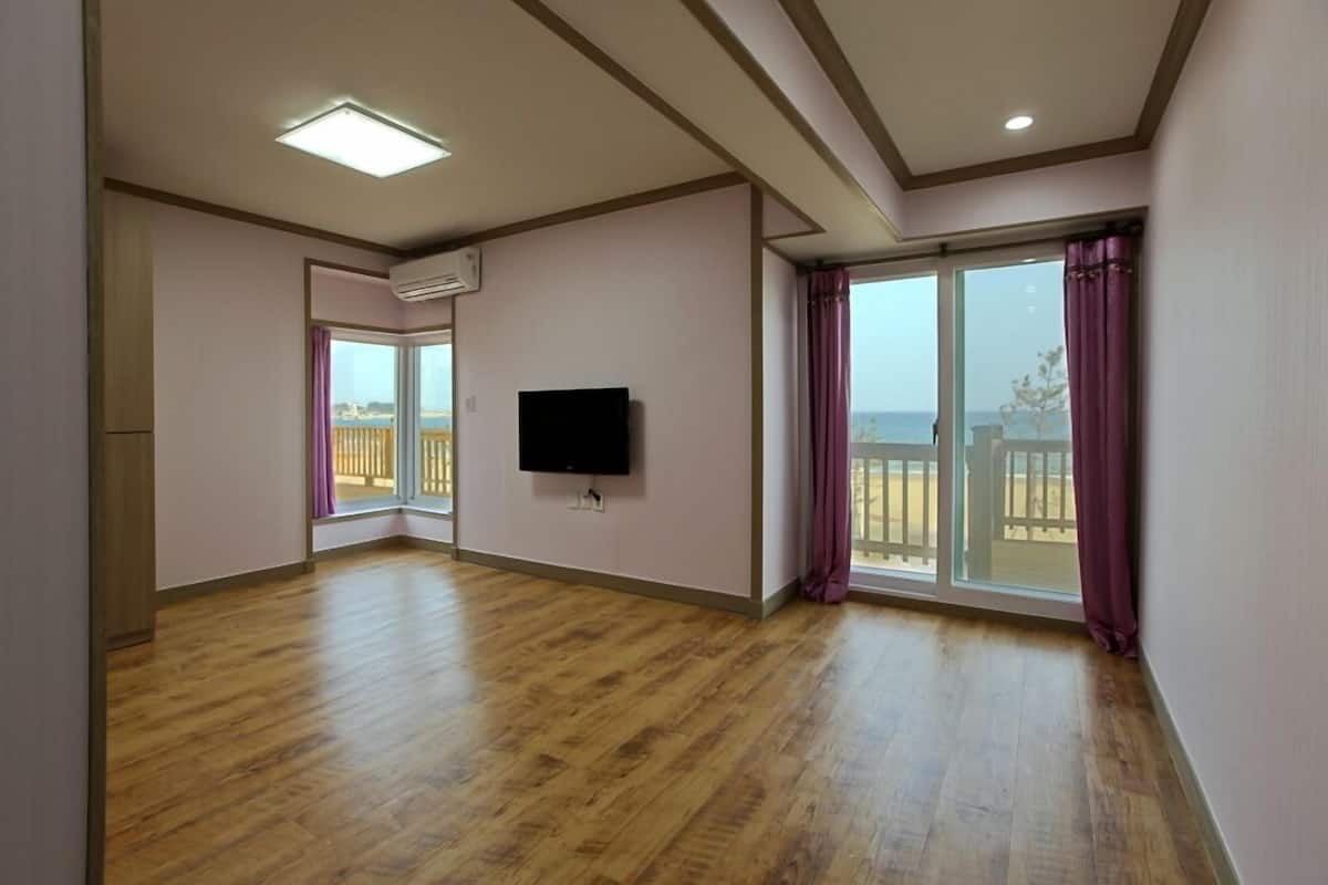 A spacious room with wooden flooring, a flat-screen TV mounted on the wall, and large sliding glass doors leading to a balcony with a view of the sea.