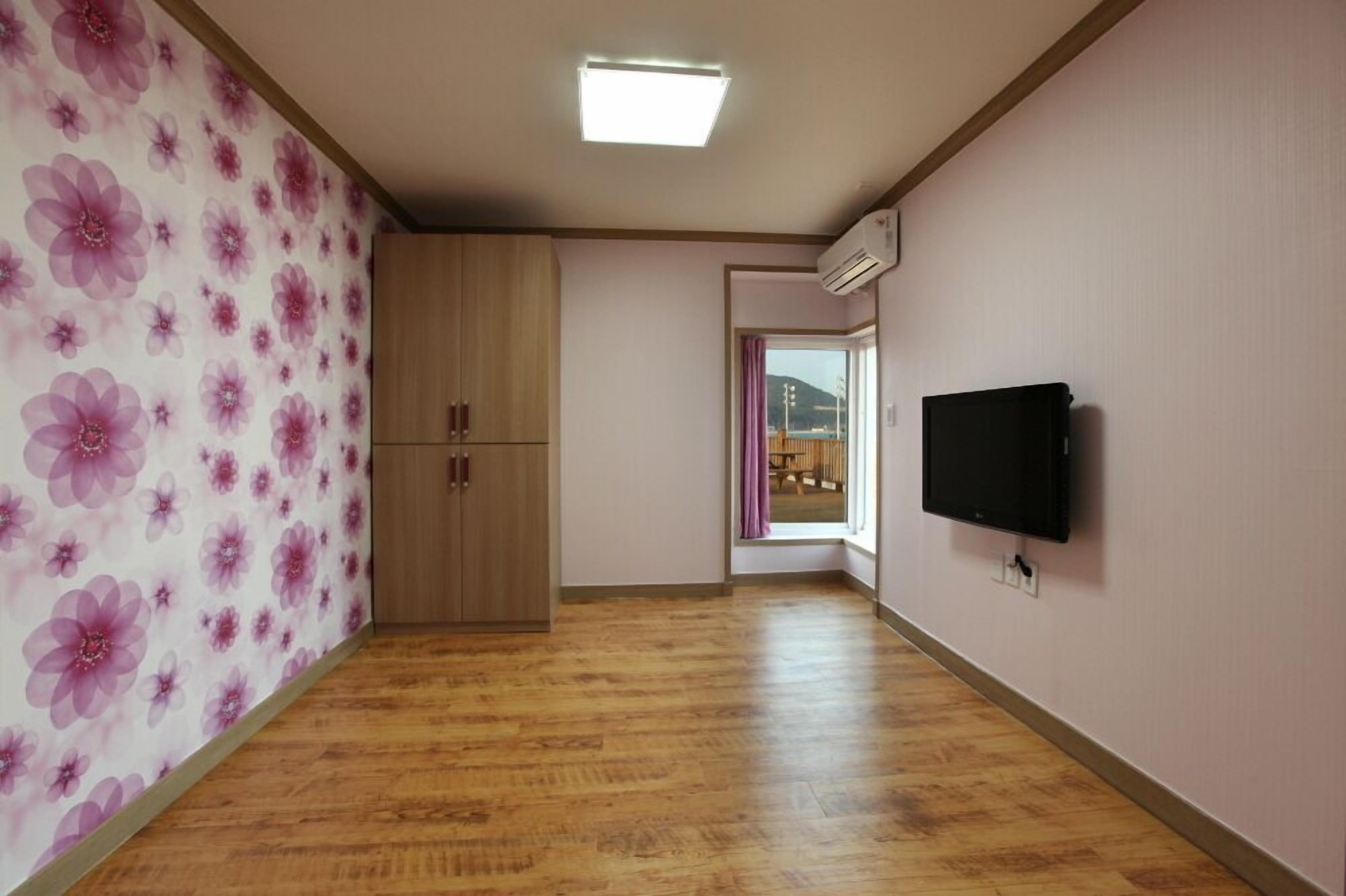 A room with wooden flooring, a wardrobe, a television, and a window with a view of a building.
