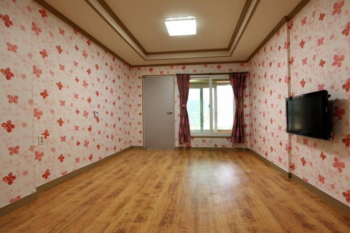 A room with floral wallpaper, a wooden floor, a door, a window with curtains, and a mounted TV.
