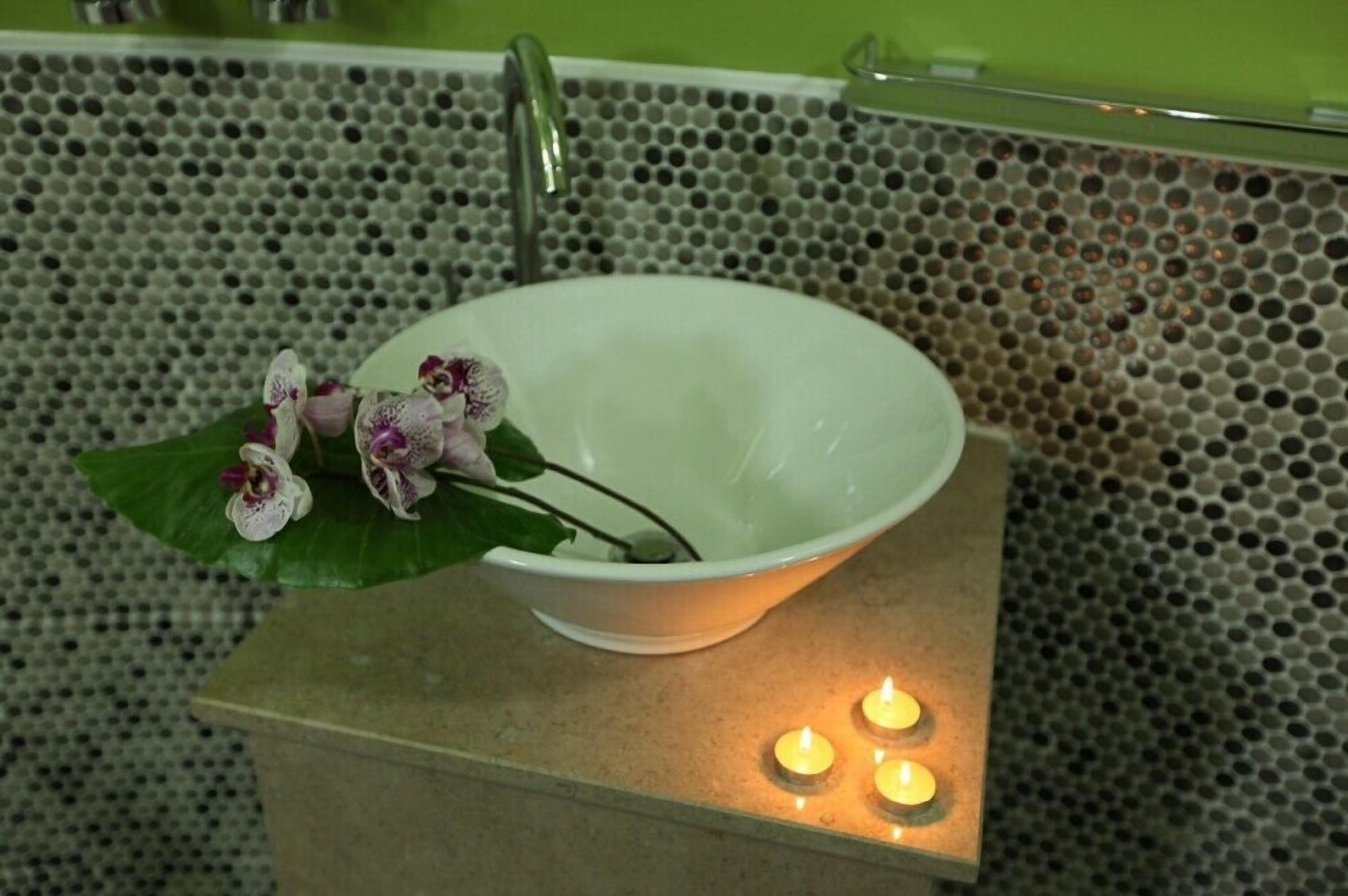 A white bowl-shaped sink with a green plant and purple flowers on a textured tile wall.