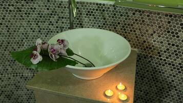 A white bowl-shaped sink with a green plant and purple flowers on a textured tile wall.