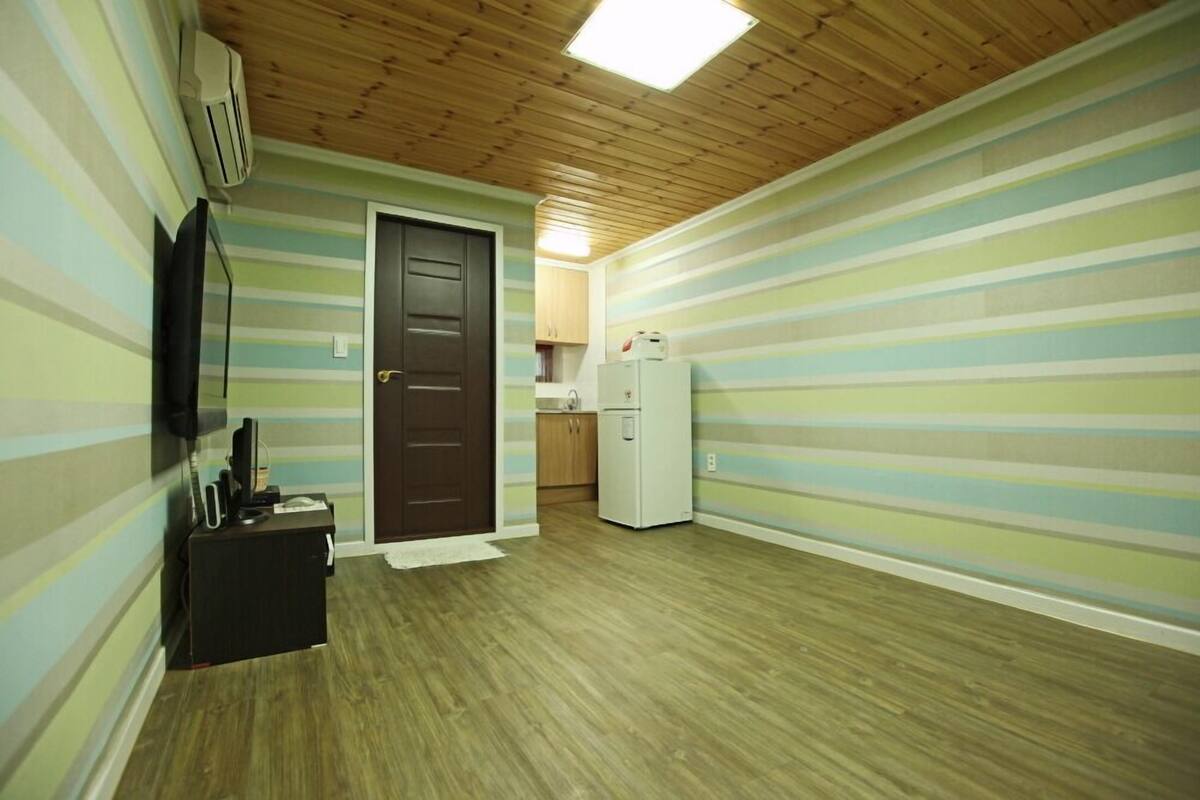 A room with striped walls, a wooden ceiling, a door, a small kitchen area, and a white refrigerator.