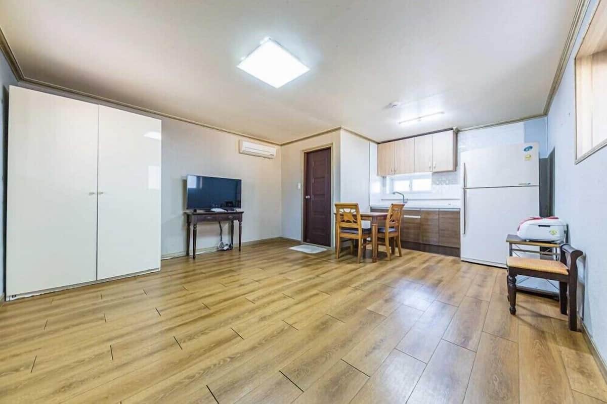 A spacious room with a wooden floor, a dining table with chairs, a television, and a kitchen area with cabinets and a refrigerator.