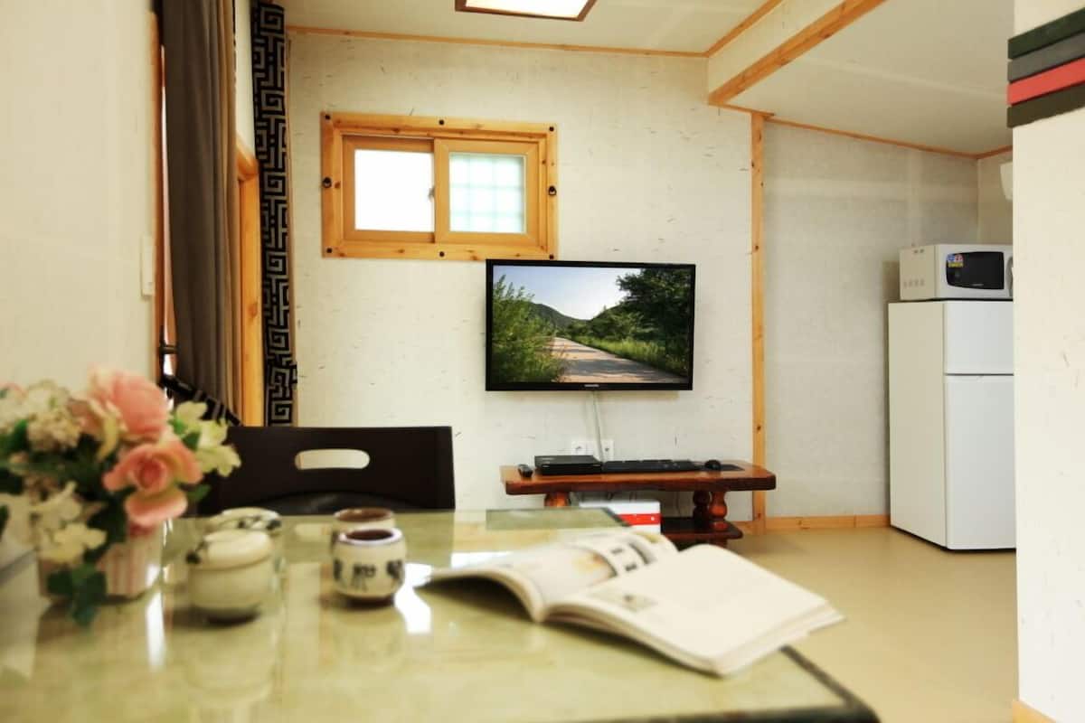 A small room with a television on the wall, a table with a book and a vase of flowers, and a refrigerator in the corner.