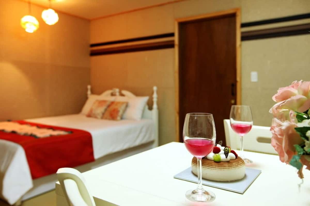 A hotel room with a bed, a dining table with two glasses of red wine, a cake, and a bouquet of flowers.