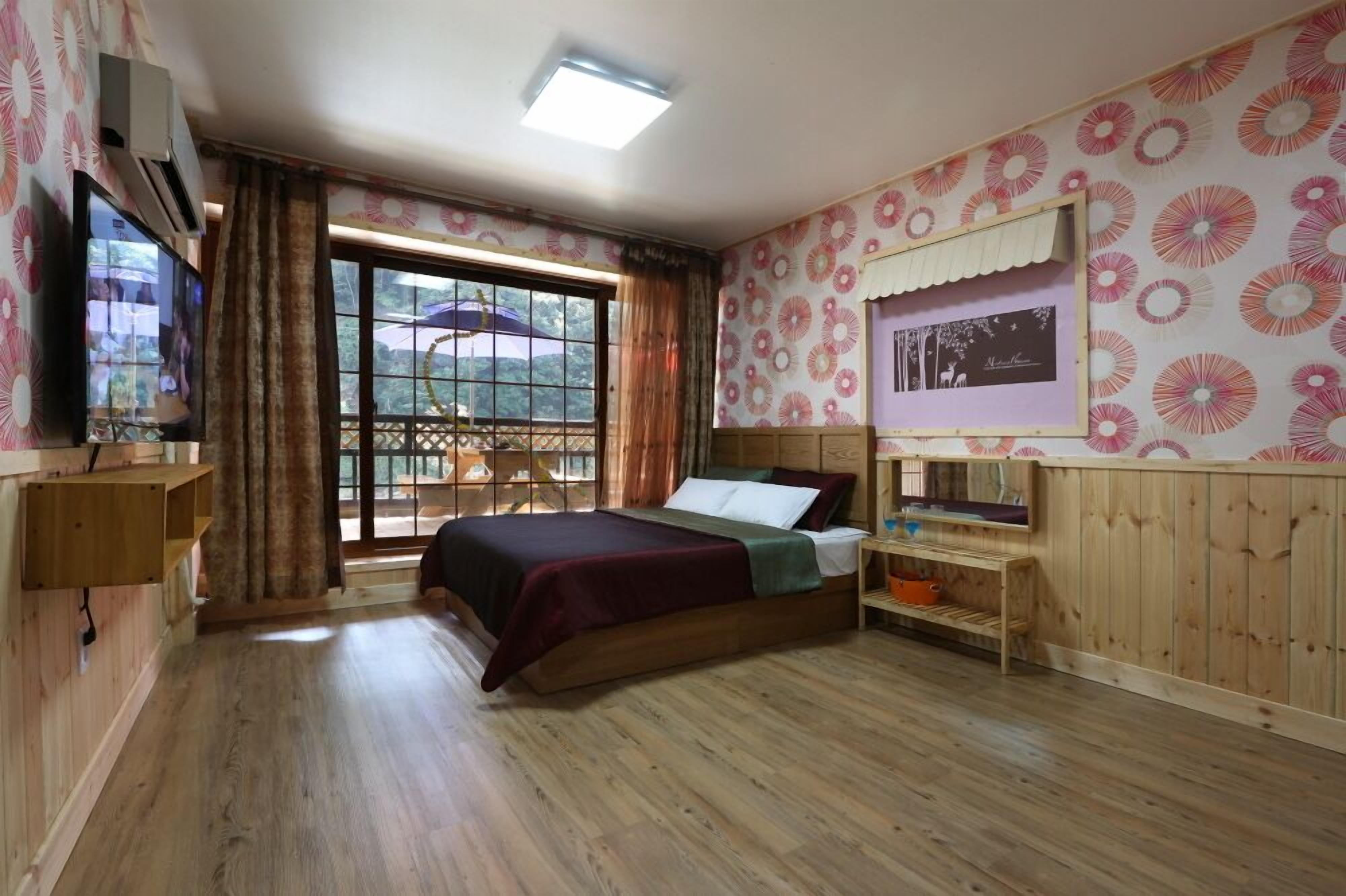 A bedroom with a wooden bed, a TV, a window with curtains, and a wall with a floral pattern.