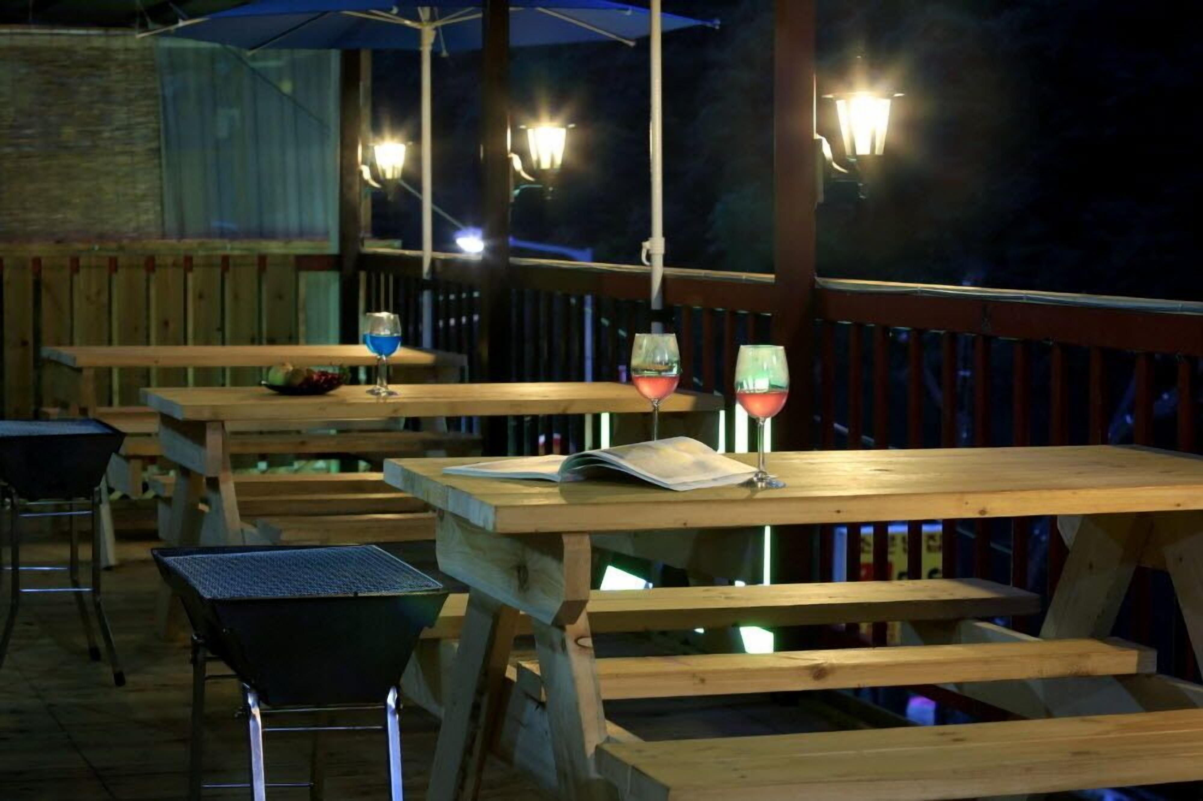 Een terras met houten tafels en banken, een barbecue en hangende lampen.