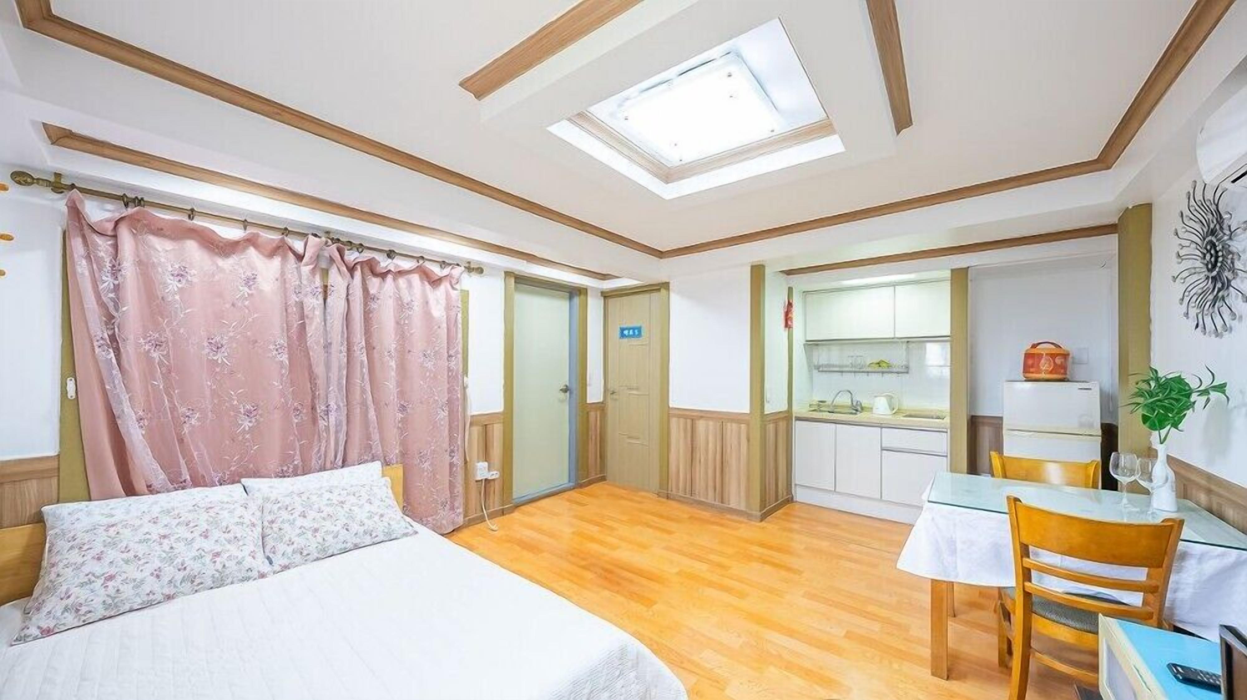 A bedroom with a bed, a small table, and a chair. There is a kitchenette in the background.
