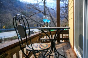 Una terraza de madera con una mesa y sillas, dos vasos de bebida azul, y vistas a árboles y una carretera.