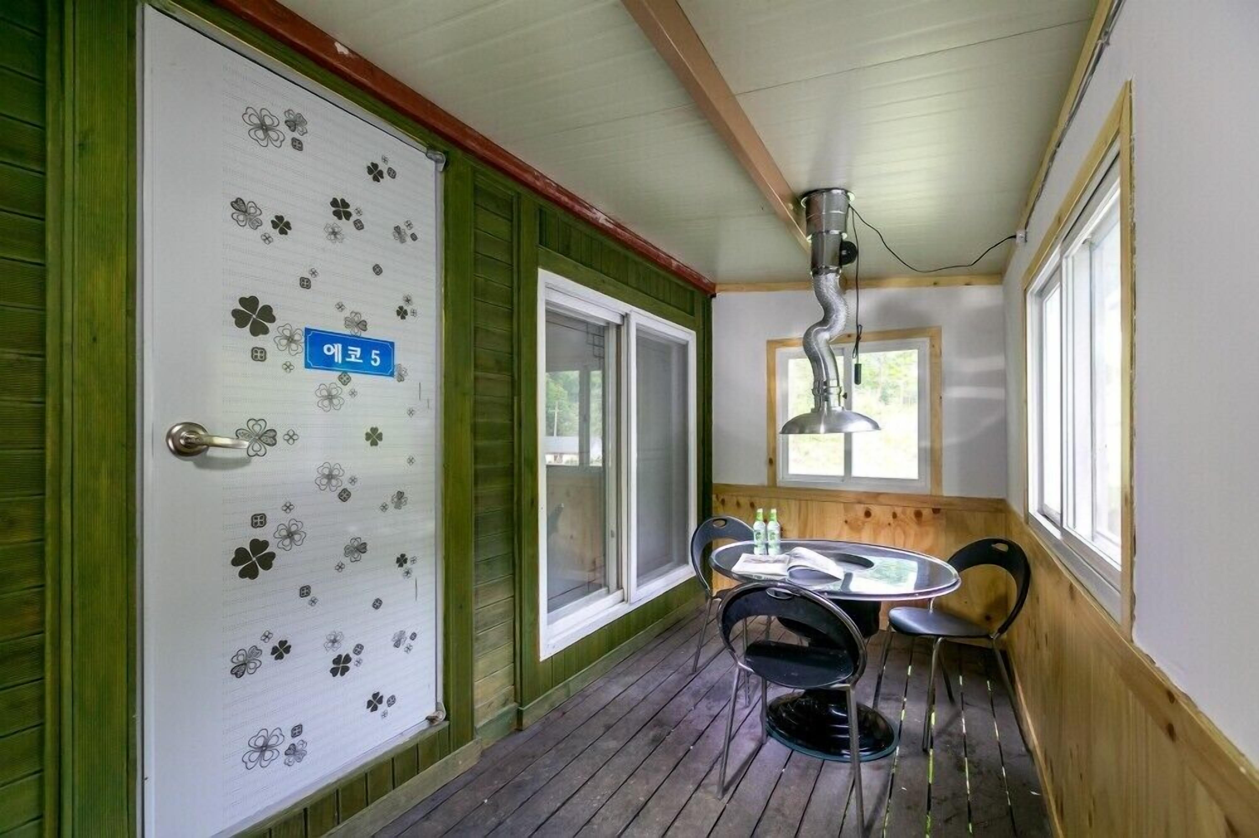 A room with a wooden floor, a table with chairs, a window, and a door with a number sign.