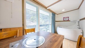 A room with a wooden dining table, a white bathtub, and a large window with a view of trees. - Eco Pia Pension (Gapyeong)